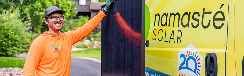 A man stands smiling holding a solar panel next to a Namaste Solar van.